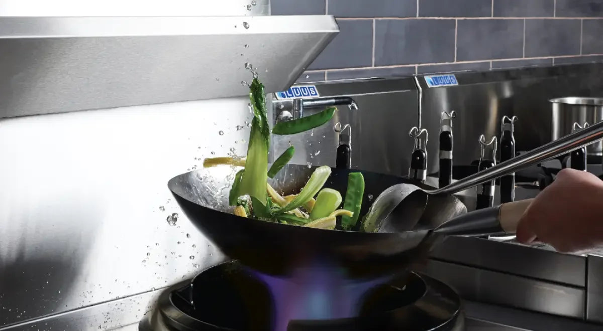 Vegetables stir-frying in a wok over a high-heat gas burner in a commercial kitchen.