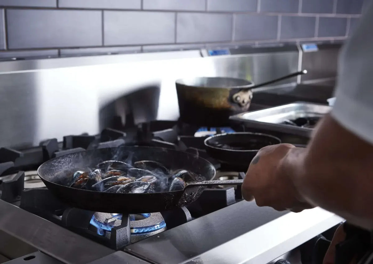 Chef cooking mussels in a frying pan on a commercial gas stove burner